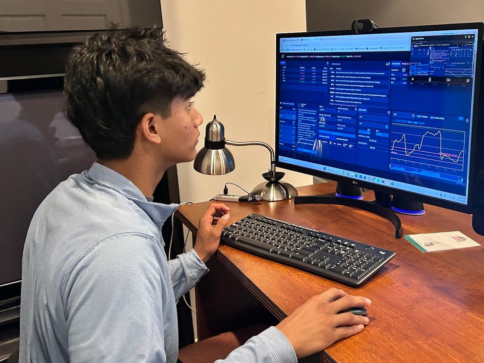 Gabriel Olasa at a desk with one hand on the mouse, watching the AmplifyME trading simulator on a desktop monitor during the CFA Institute Investing Simulator Challenge