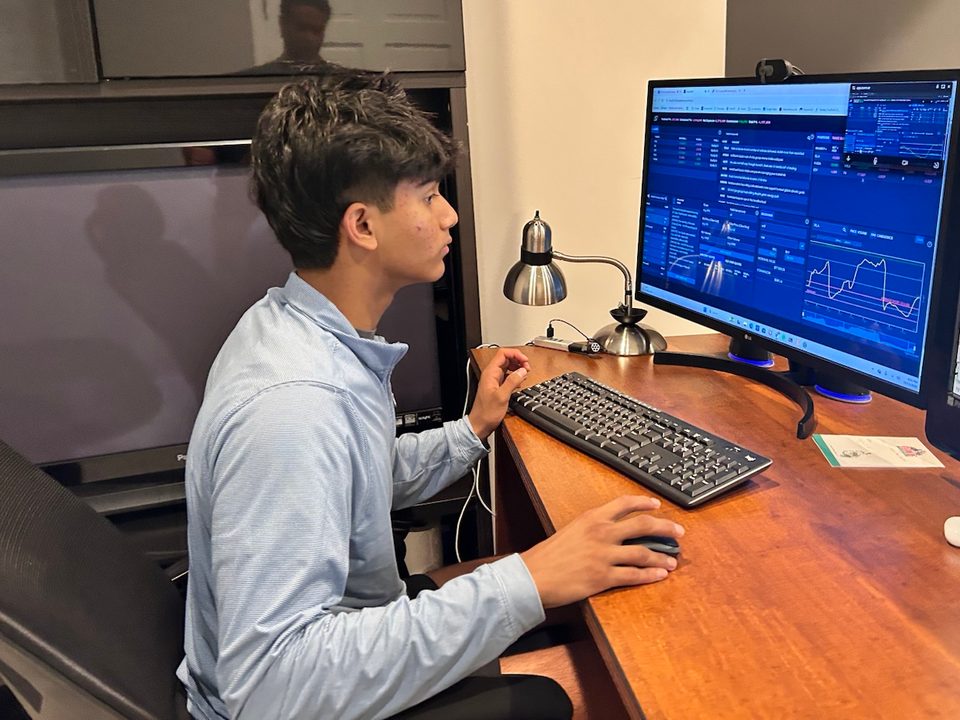 Gabriel Olasa at a desk with one hand on the mouse, watching the AmplifyME trading simulator on a desktop monitor during the challenge.